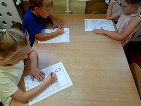 Troje dzieci siedzi przy stoliku i piszę ołówkami zera na kartach papieru A4.
