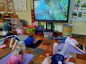 Dzieci leżą na matach i spoglądają na monitor, na którym wyświetlone jest zdjęcie drzew liściastych. 