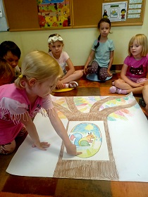 Dzieci siedzą na podłodze wokół plakatu z drzewem, na którym leży rysunek dziupli z wiewiórką. Jedna dziewczynka wskazuje palcem dziuplę z wiewiórką. 
