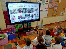 Dzieci siedzą przed monitorem na podłodze, na poduszkach. Na monitorze wyświetlane są zdjęcia, które przedstawiają okulistów. 