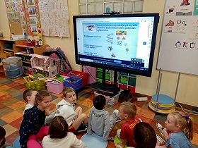 Dzieci siedzą przed monitorem na podłodze, na poduszkach. Na monitorze wyświetlane jest zdjęcie, które pokazuje za co odpowiada zmysł wzroku.