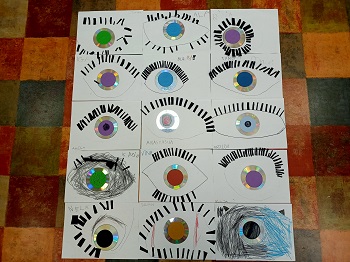 Na podłodze leżą prace plastyczne dzieci, które ukazują oko. Prace wykonane są z płyt CD, z kolorowym środkiem oraz z małych, czarnych karteczek. 