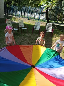 Troje dzieci - 2 dziewczynki i jeden chłopiec trzymają kolorowa chustę animacyjną. Stoją na trawniku. Za nimi stoją 4 krzesła. 