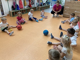 Dzieci siedzą w dwóch rzędach naprzeciw siebie i turlają do siebie piłkę czerwoną i niebieską.