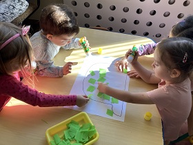Przy stoliku siedzi czworo dzieci, przyklejają zielone kawałki krepiny wypełniając narysowany kształt.
