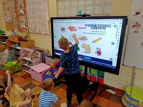 Chłopiec stoi przed monitorem i zakreśla obrazki, które rozpoczynają się na głoskę o. Reszta dzieci siedzi na podłodze i patrzy na to, co robi chłopiec. 