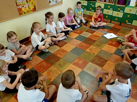 Dzieci siedzą w siadzie skrzyżnym na podłodze, w strojach gimnastycznych i głaszczą się po dłoniach.