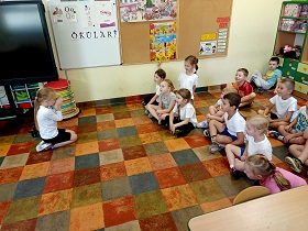 Dzieci siedzą w siadzie skrzyżnym na podłodze, w strojach gimnastycznych i obserwują dziewczynkę, która klęczy i składa ręce przy nosie.