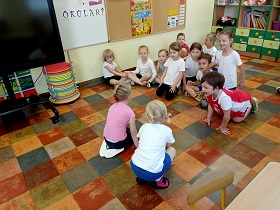 Dzieci siedzą w siadzie skrzyżnym na podłodze, w strojach gimnastycznych i obserwują dwie dziewczynki, która klęczą i im coś pokazują.