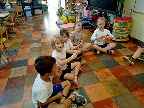 Dzieci siedzą w siadzie skrzyżnym na podłodze, w strojach gimnastycznych i składają ręce, pokazując coś. 