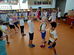 Dzieci w strojach gimnastycznych bawią się wspólnie. Jedna dziewczynka z zieloną szarfą, przekłada hula hop przez chłopca, który trzyma ręce do góry.