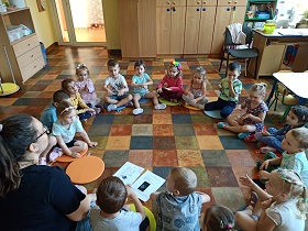 Dzieci oraz nauczycielka siedzą w sali w kole. Wszyscy trzymają ręce przed sobą i głaszczą swoje dłonie w ramach zabawy paluszkowej. 