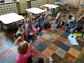 Dzieci siedzą w sali w kole. Wszyscy mają podniesioną prawą rękę i dłoń położoną przy czole. Pokazują "cześć" w języku migowym.