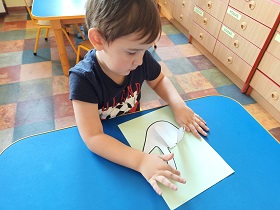 Przy stoliku siedzi chłopiec. Przed nim leży wykonana przez niego praca -biala dłoń przyklejona do zielonej kartki. Chłopiec zagina palce aby wskazywały napis love w języku migowym.