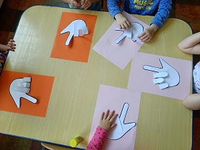 Na stole leży 5 prac dzieci - 3 na różowych kartkach i 2 na pomarańczowych. Na kartkach przyklejone są dłonie wycięte z papieru pokazujące znak "love" w języku migowym. Palce mały, serdeczny i środkowy są zagięte do środka. 