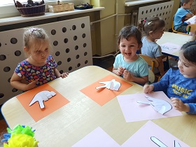 Przy stoliku siedzą 3 dziewczynki. Przed sobą mają położone swoje prace przedstawiające napis "love". W tle widać inne dzieci siedzące przy stolikach. 