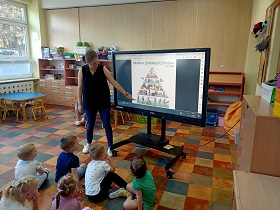 Dzieci siedzą na podłodze przed monitorem, przy którym stoi pani i wskazuje palcem na wyświetlane zdjęcie piramidy zdrowego żywienia dla dzieci.