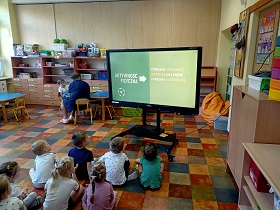 Dzieci siedzą na podłodze przed monitorem, na którym wyświetlane jest zdjęcie na zielonym tle. Na zdjęciu napis: aktywność fizyczna. Z tyłu za monitorem siedzi pani z chłopcem.