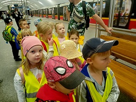 Dzieci stoją w parach na peronie stacji metra w żółtych, odblaskowych kamizelkach. 