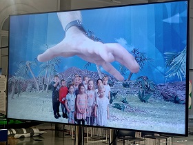 Na ekranie telewizora widać dzieci, które stoją w grupie. Nad nimi znajduje się wielka dłoń, która chce je złapać.
