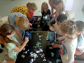 Dzieci stoją i siedzą nad tabletami, które ustawione są na stojakach. Pod tabletami leżą papierowe rysunki, które dzieci kładą pod tabletem, by zrobić im zdjęcie.