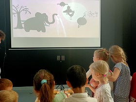 Dzieci stoją nad podświetlonym ekranem, po którym przesuwają kostkami. Patrzą na ekran zawieszony na ścianie, na którym wyświetlana jest animacja, którą tworzą kostki.
