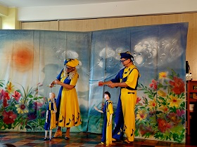 Pan i Pani przebrani w stroje teatralne, żółto-niebieskie, trzymają w dłoniach marionetki i nimi grają. Za nimi stoi kurtyna.