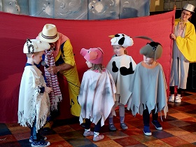 Czwórka dzieci w przebraniach zwierząt stoi przed czerwoną kurtyną, którą trzyma pani w kapeluszu. Obok chłopca w przebraniu barana stoi pan, który podaje mu mikrofon.
