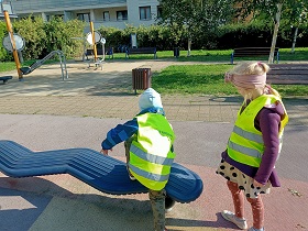 Dziewczynka i chłopiec w odblaskowych kamizelkach stoją przy ławeczce do ćwiczenia utrzymywania równowagi. Chłopiec na nią wchodzi. 