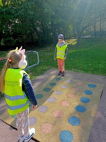 Dziewczynka i chłopiec w odblaskowych kamizelkach stoją przy planszy zrobionej na terenie placu zabaw, która przedstawia kolorowe kółka.