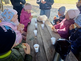 Dzieci siedzą przy drewnianym stole i jedzą bułki. Na stole stoją plastikowe kubeczki z herbatą oraz termos. Obok dzieci stoją dorośli. 