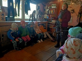 Dzieci siedzą w pomieszczeniu na ławkach i obserwują panią w fioletowej kurtce i zielonej czapce, która trzyma w rękach uprząż dla konia.