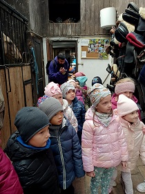 W stajni stoi grupa dzieci, która jest ubrana w kurtki i czapki. Za nimi stoi pan w fioletowej bluzie.