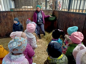 Dzieci wraz z panią w fioletowej kurtce i czapce, stoją na sianie w stajni. Dzieci są zwrócone twarzami do pani. 
