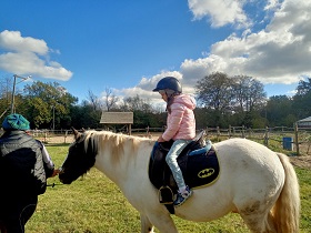 Dziewczynka w różowej kurtce i toczku, siedzi na koniu, którego prowadzi pani. 