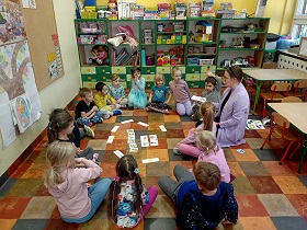 Dzieci siedzą w siadzie skrzyżnym w kółeczku. Na środku kółka leżą ilustracje i napisy. Za dziećmi znajdują się szafki z zabawkami.