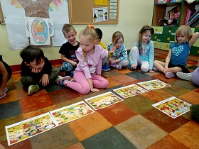 Dziewczynka w różowym stroju patrzy na ilustracje, które leżą w jednym rzędzie. Za nią siedza inne dzieci, które spoglądają na to, co ona robi.