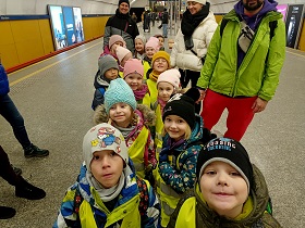 Dzieci w żółtych, odblaskowych kamizelkach stoją na peronie w metrze. Ustawione są w parach. Razem z nimi stoją opiekunowie.