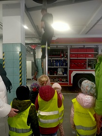 Dzieci w żółtych, odblaskowych kamizelkach obserwują strażaka, który zjeżdża po rurze. Za nim stoi wóz strażacki.