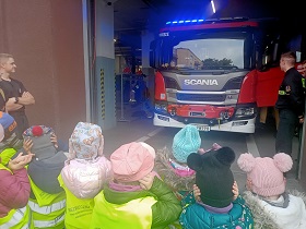 Dzieci w żółtych, odblaskowych kamizelkach zatykają uszy. Przed nimi stoi wóz strażacki, który puszcza sygnał dźwiękowy, w związku z tym palą się niebieskie światła. 
