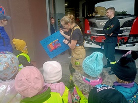 Dzieci w żółtych, odblaskowych kamizelkach stoją przed panią, kora otrzymała od chłopca prezent w postaci wyklejonego bibułą wozu strażackiego. 