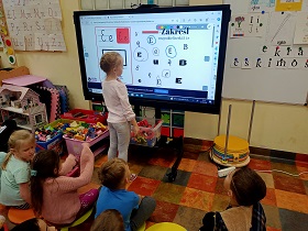 Dziewczynka stoi przy monitorze i zaznacza na nim literkę e w kółku. Reszta dzieci siedzi na podłodze i na nią spogląda. 