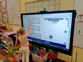 Dziewczynka stoi przy monitorze i kreśli linie w labiryncie w literce e. 
