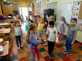 Dzieci stoją w sali i podnoszą do góry prawą rękę, na której mają założoną szarfę. 