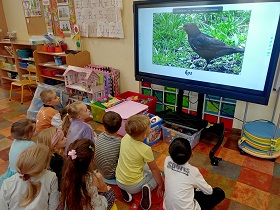 Dzieci siedzą na podłodze przed monitorem, na którym wyświetlany jest film przedstawiający kosa.