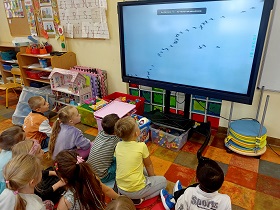 Dzieci siedzą na podłodze przed monitorem, na którym wyświetlany jest film przedstawiający lecące w kluczu żurawie. 