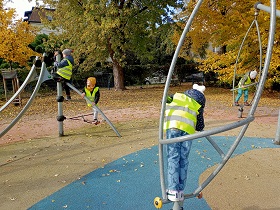 Dzieci w odblaskowych kamizelkach bawią się na placu zabaw wchodząc na sprzęty. 