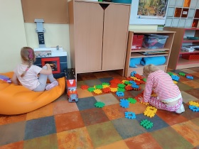 Jedna dziewczynka siedzi na dużej poduszce i bawi przy przy zabawkowej kuchence, druga dziewczynka układa duże klocki na podłodze