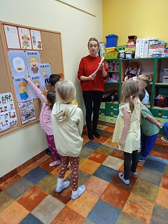 Dzieci wraz z panią w czerwonej bluzce stoją przy korkowej tablicy i wskazują na kartkach emocje. 