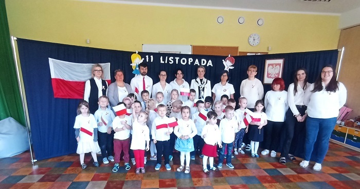 Dzieci i dorośli ubrani w stroje galowe stoją na baczność. Za nimi stoi granatowa kotara, na której widoczna jest biało-czerwona flaga, godło Polski, dwoje postaci - chłopiec i dziewczynka oraz napis 11 listopada. Dzieci trzymają w dłoniach biało-czerwone flagi. 